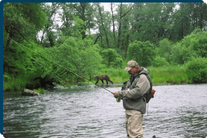 A man fishing in the river with a bear on the shore.